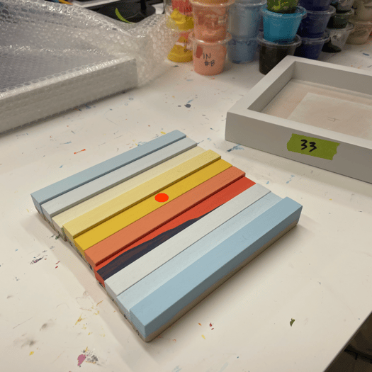 photo of an incomplete wooden slat artwork in blues, yellows, oranges showing a sunset