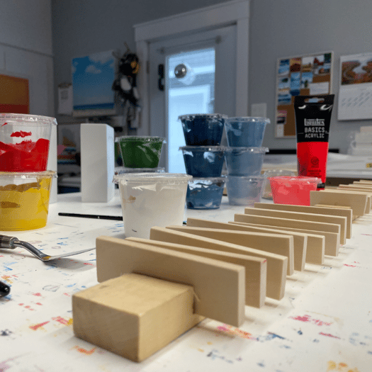 Photo of unpainted wooden slats on a studio table surrounded by small containers of bright paints